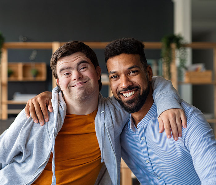 Happy young man with Down syndrome and his tutor with arms around looking at camera indoors at school